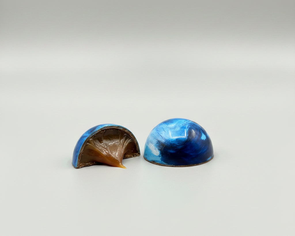 a milk chocolate sea salt creme caramel hand painted in multiple blue colored cocoa butters symbolizing waves and shown next to its cut-open half revealing sea salt creme caramel inside.